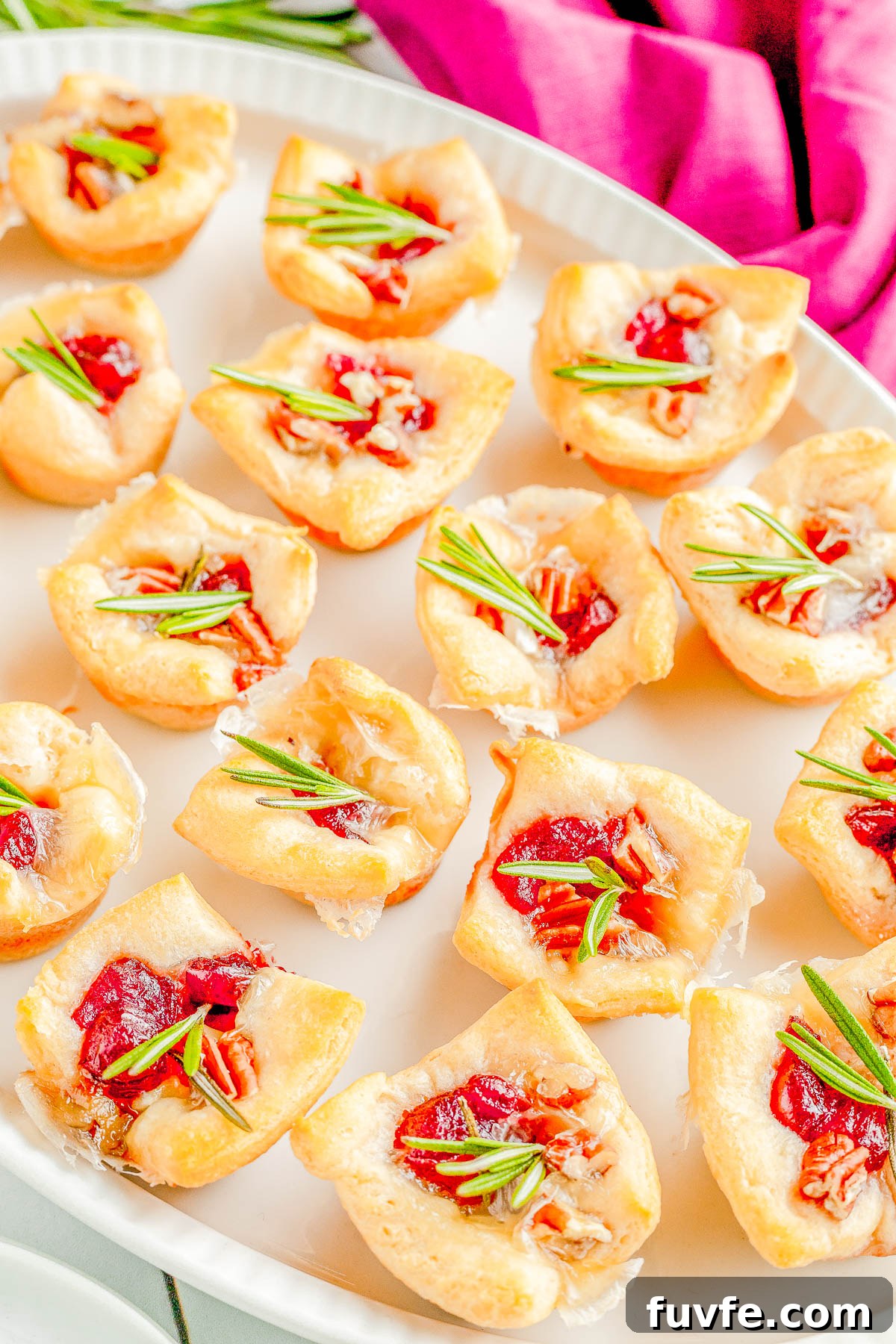 A platter of baked cranberry brie bites topped with chopped pecans and fresh rosemary. 