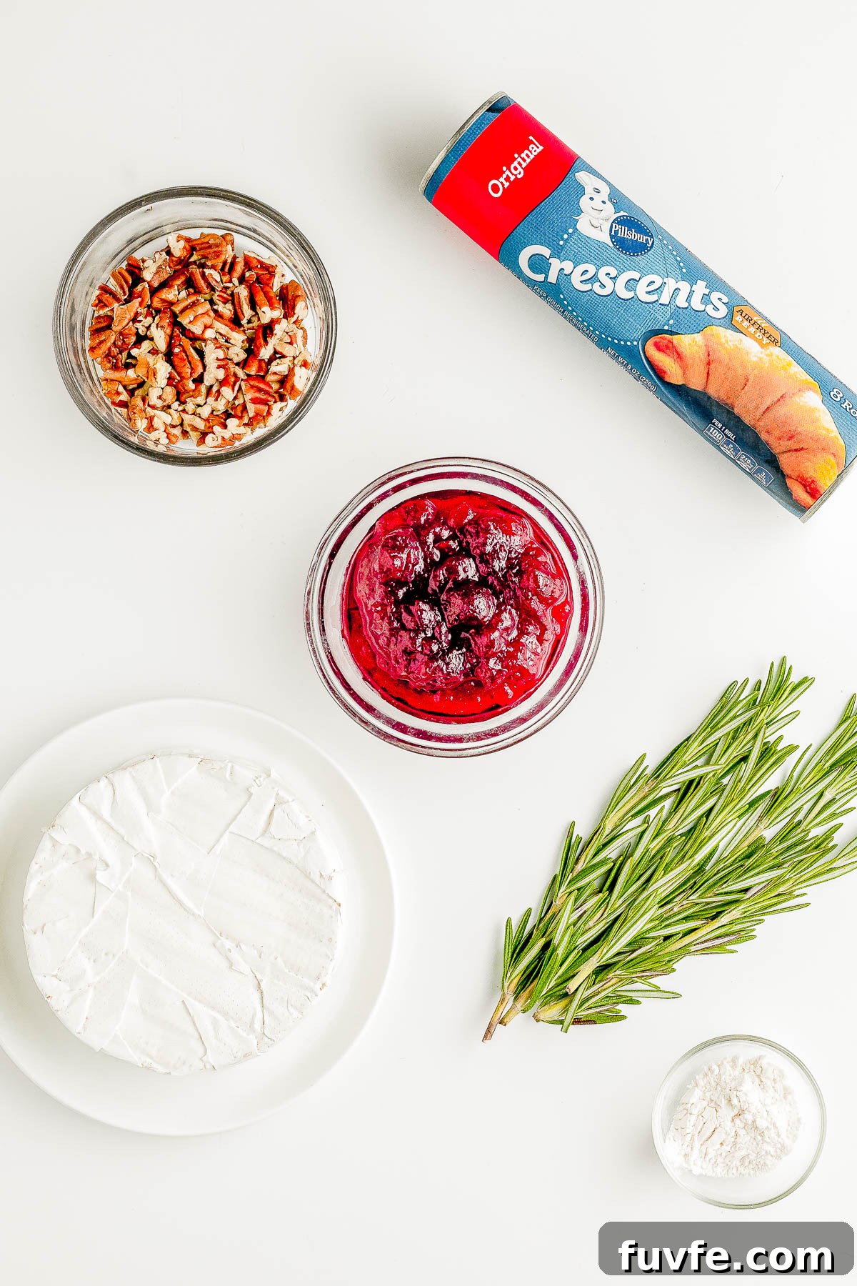 Brie, fresh rosemary, a glass bowl of cranberry sauce, a glass bowl of chopped pecans, and a package of crescent roll dough. 