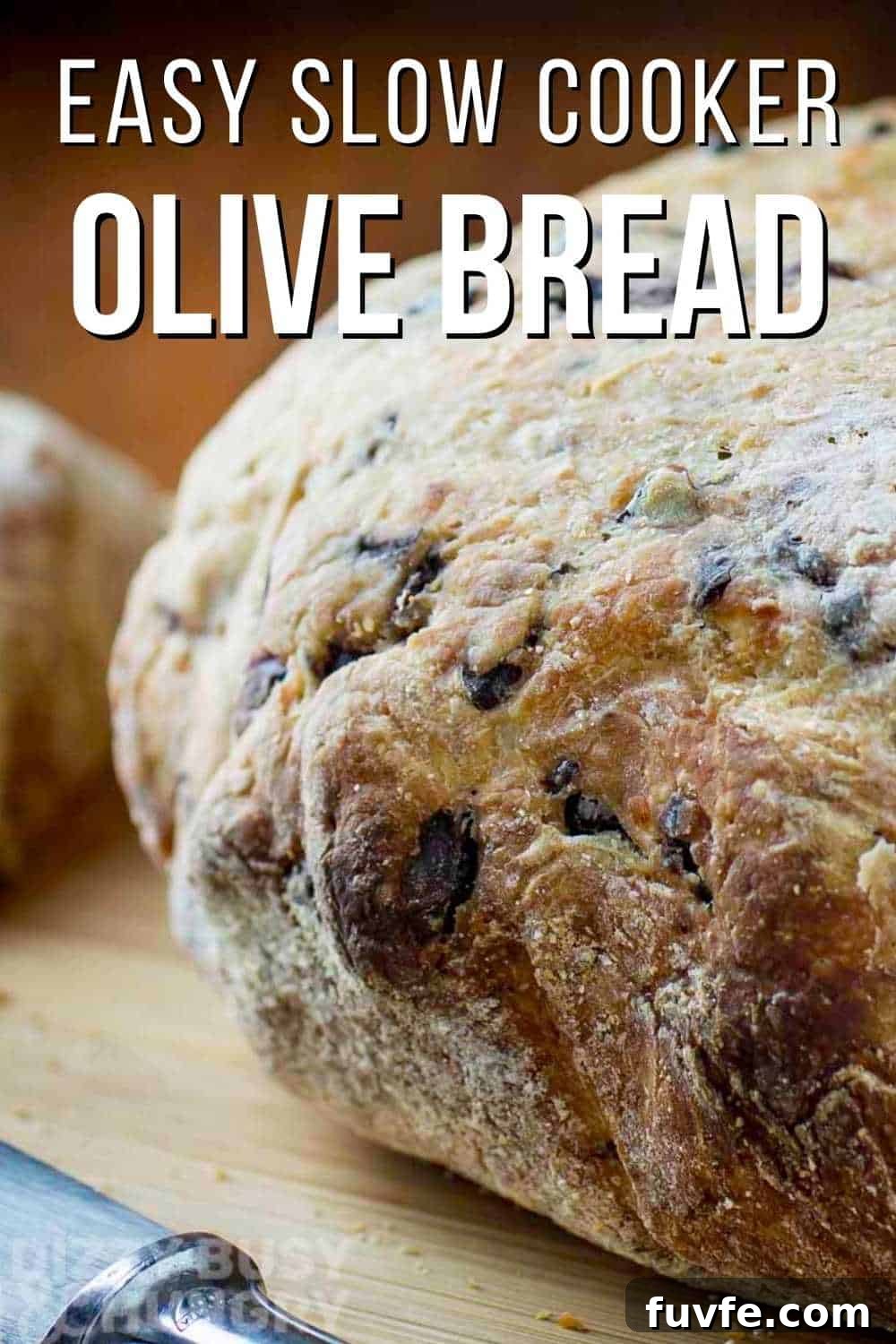 Side view of whole loaf of olive bread on a wooden cutting board with a butter knife on the side.