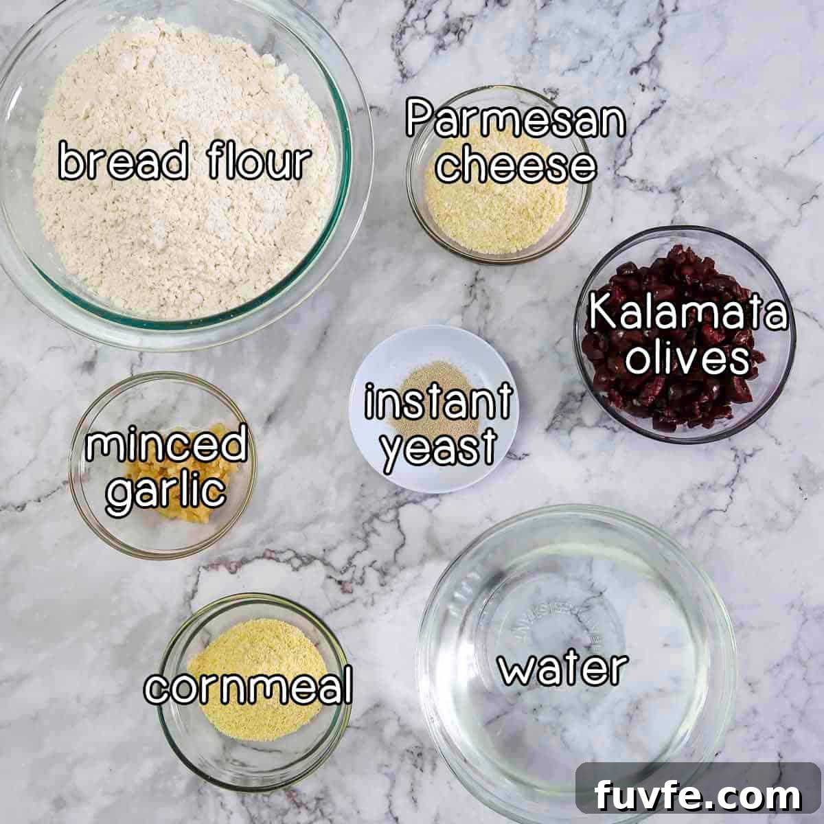 Overhead shot of ingredients- bread flour, parmesan cheese, Kalamata olives, instant yeast, minced garlic, cornmeal, and water.