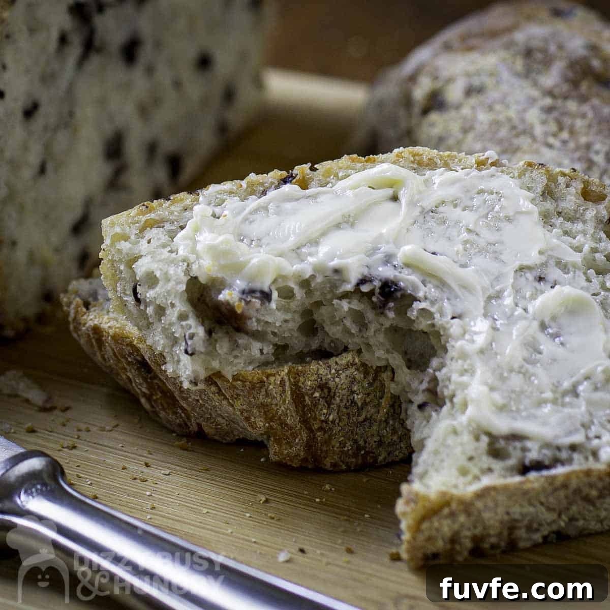 Close up shot of slice of olive bread with butter with whole loaf in the background on a cutting board with a butter knife in front.