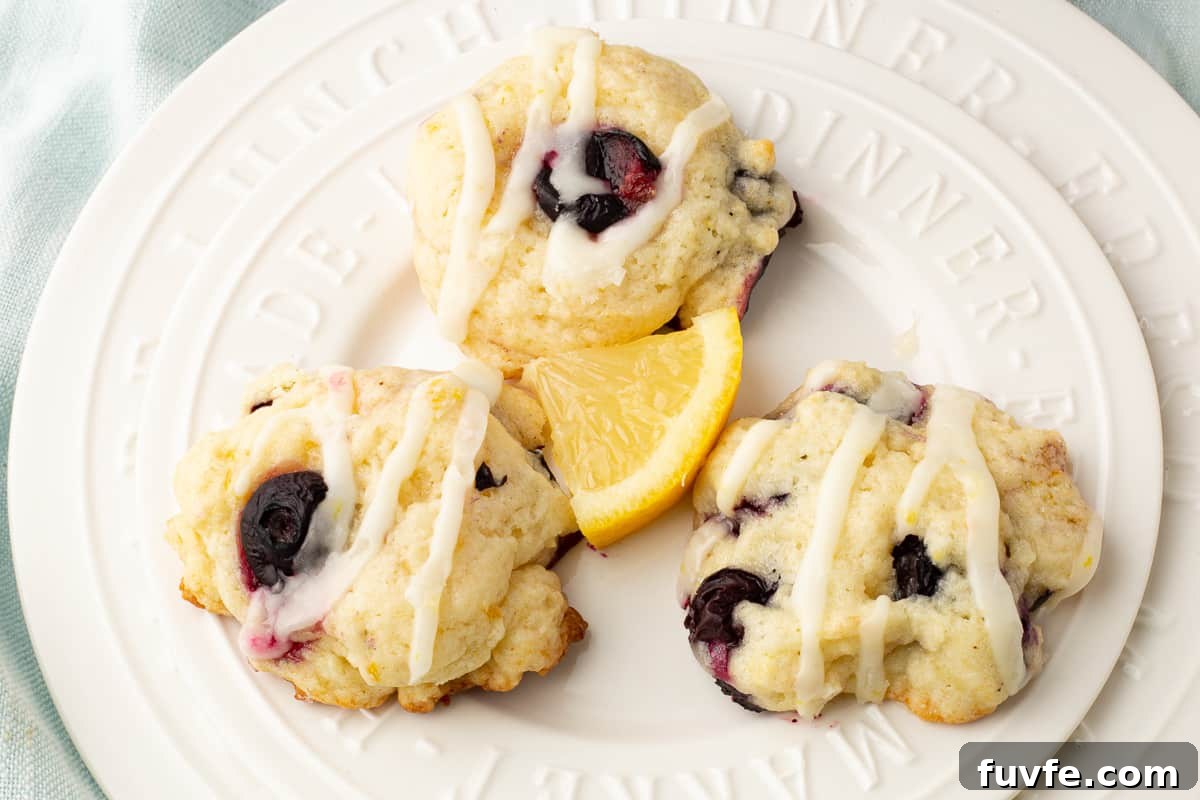 Soft Lemon Blueberry Cookies Recipe: Tender Citrus Berry Treats 9 Three glaze lemon blueberry cookies on a white plate with a slice of lemon.