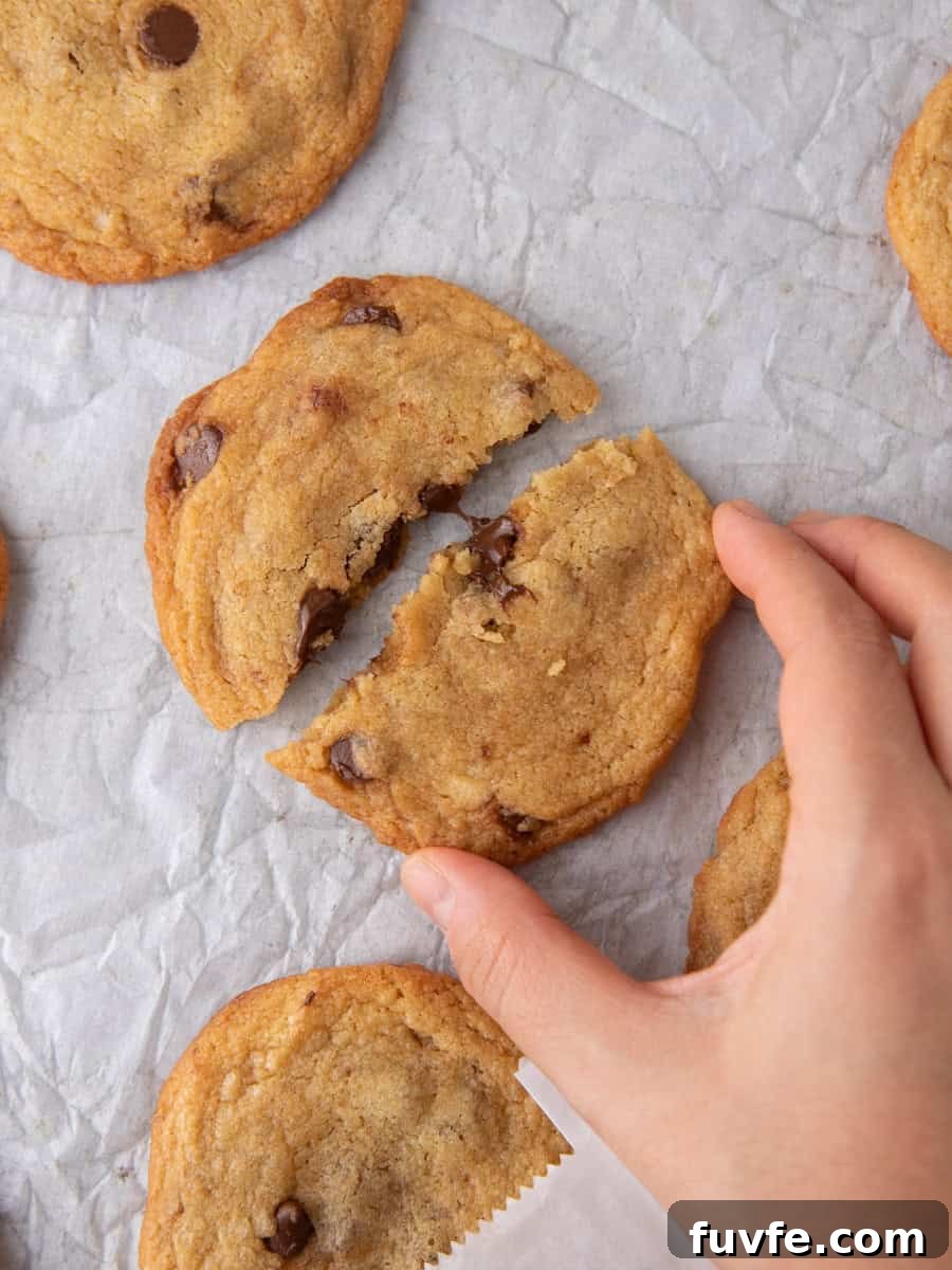 Hand breaking a freshly baked thin chocolate chip cookie showing chewy center.