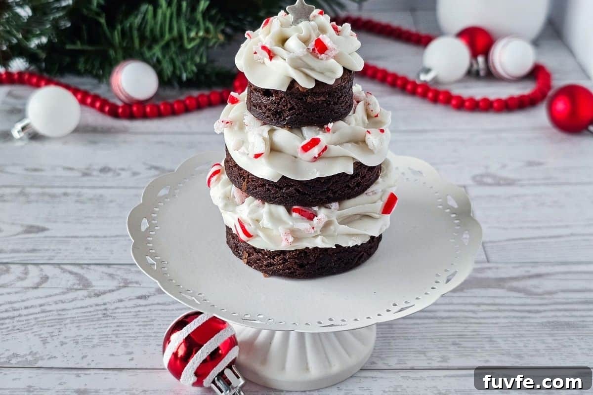 29 Christmas Baking Ideas for Home Bakers Who Love a Busy Oven 2 A stack of three round chocolate brownies with white frosting and crushed peppermint, arranged in a tree shape on a white cake stand with holiday decorations in the background.