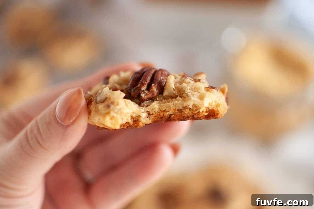 29 Christmas Baking Ideas for Home Bakers Who Love a Busy Oven 14 A hand holds a bitten Butter Pecan Cookie topped with a pecan and chopped nuts, with a blurred background.