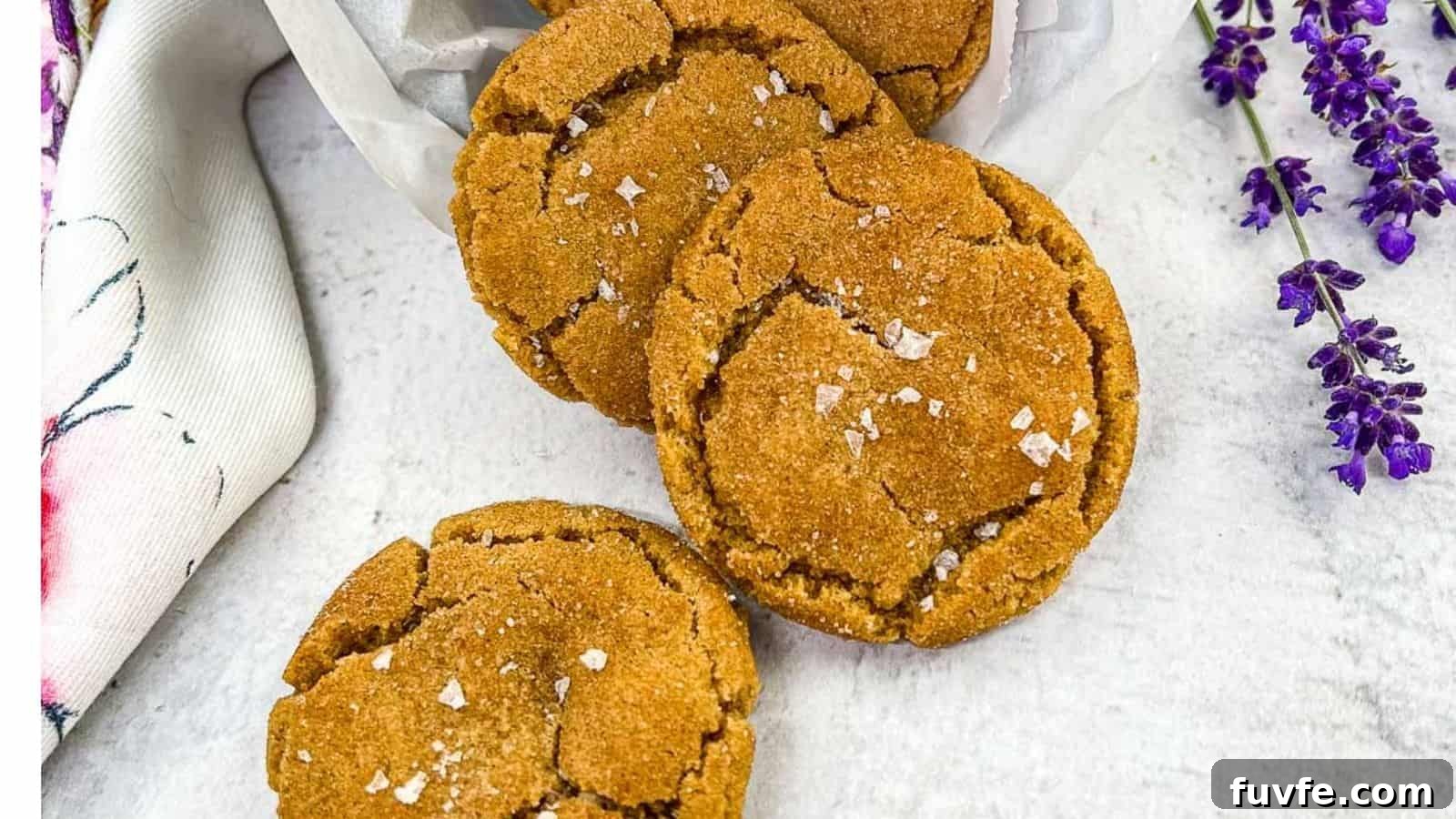 29 Christmas Baking Ideas for Home Bakers Who Love a Busy Oven 31 Three brown butter snickerdoodle cookies arranged on a white surface, sprinkled with sea salt flakes, with a white cloth and purple flowers on the side.