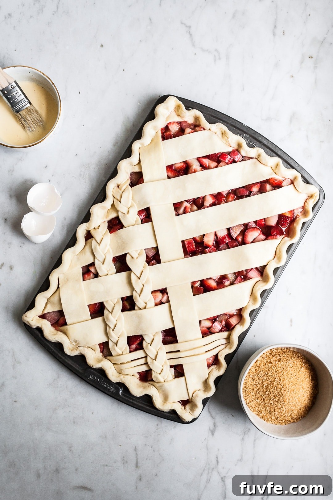 Strawberry Rhubarb Slab Pie Recipe — Perfect for Parties 5 Crimped unbaked lattice slab pie crust in sheet pan on a marble background with bowls of sugar and egg wash nearby