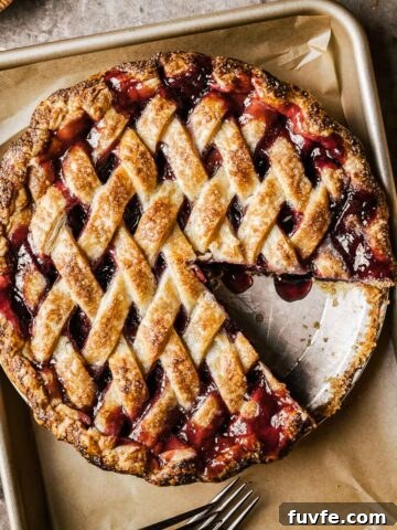 A golden brown lattice topped baked cherry rhubarb pie on a parchment lined baking sheet. A slice has been removed from the pie.