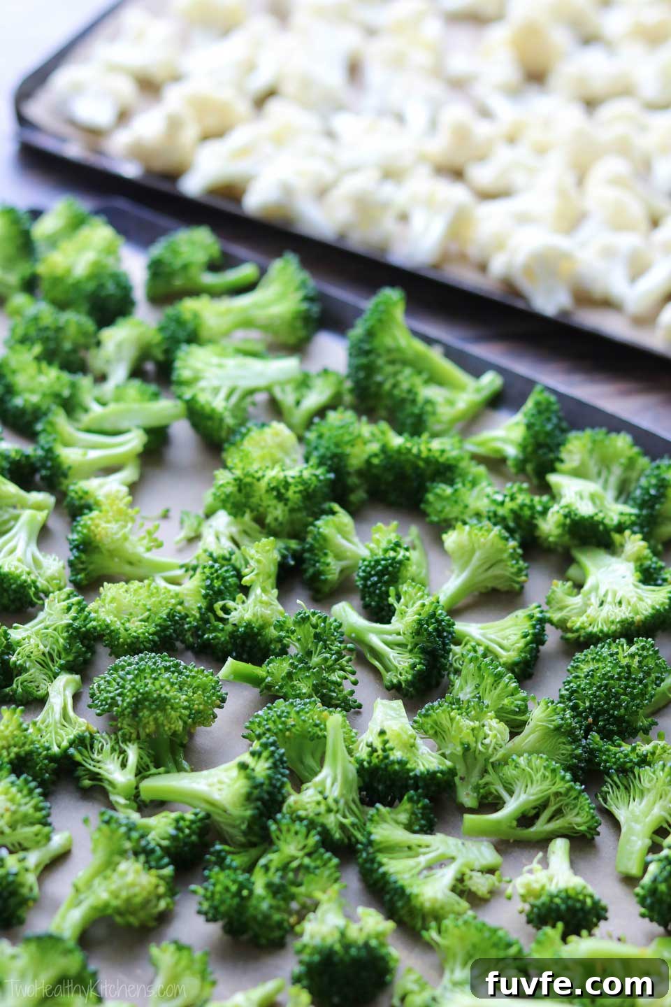 two sheet pans, ready to go into the oven for roasting - one in the foreground with broccoli spread on it, and the other in the background with cauliflower