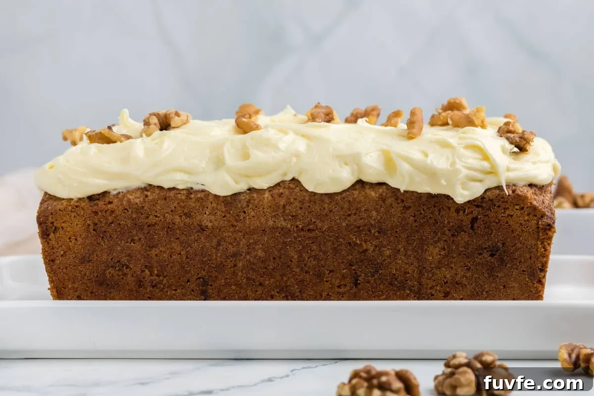 A loaf of carrot cake topped with cream cheese frosting and walnut halves sits on a white rectangular plate.
