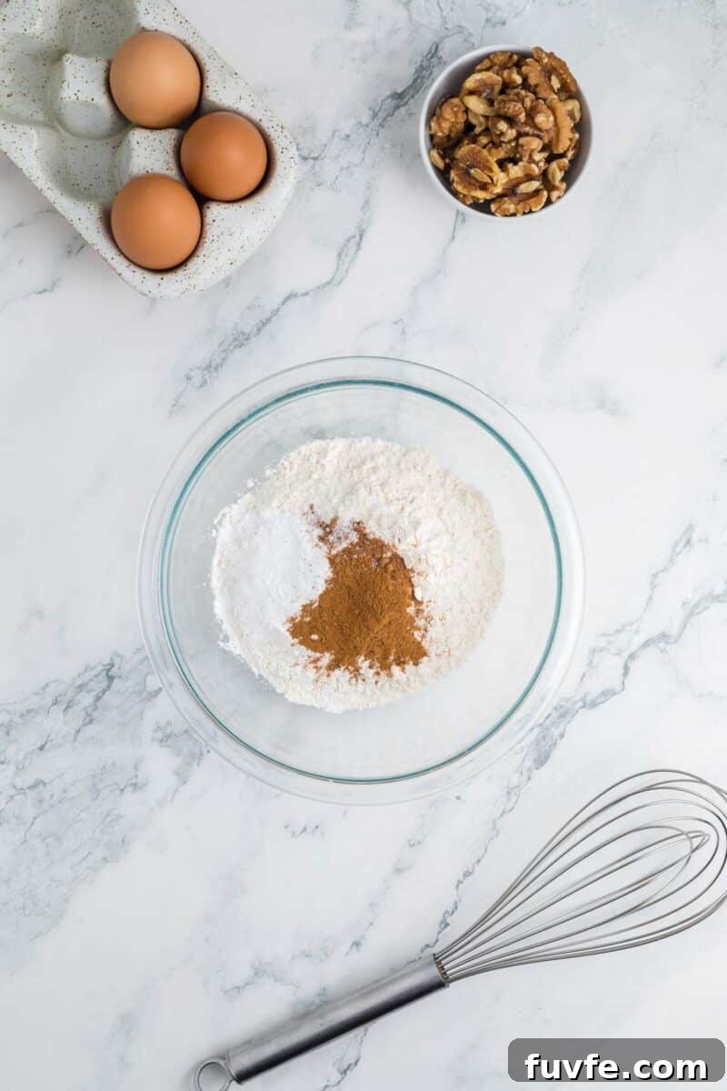 A glass bowl with flour, baking powder, and cinnamon on a marble surface, with eggs, walnuts, and a metal whisk nearby.