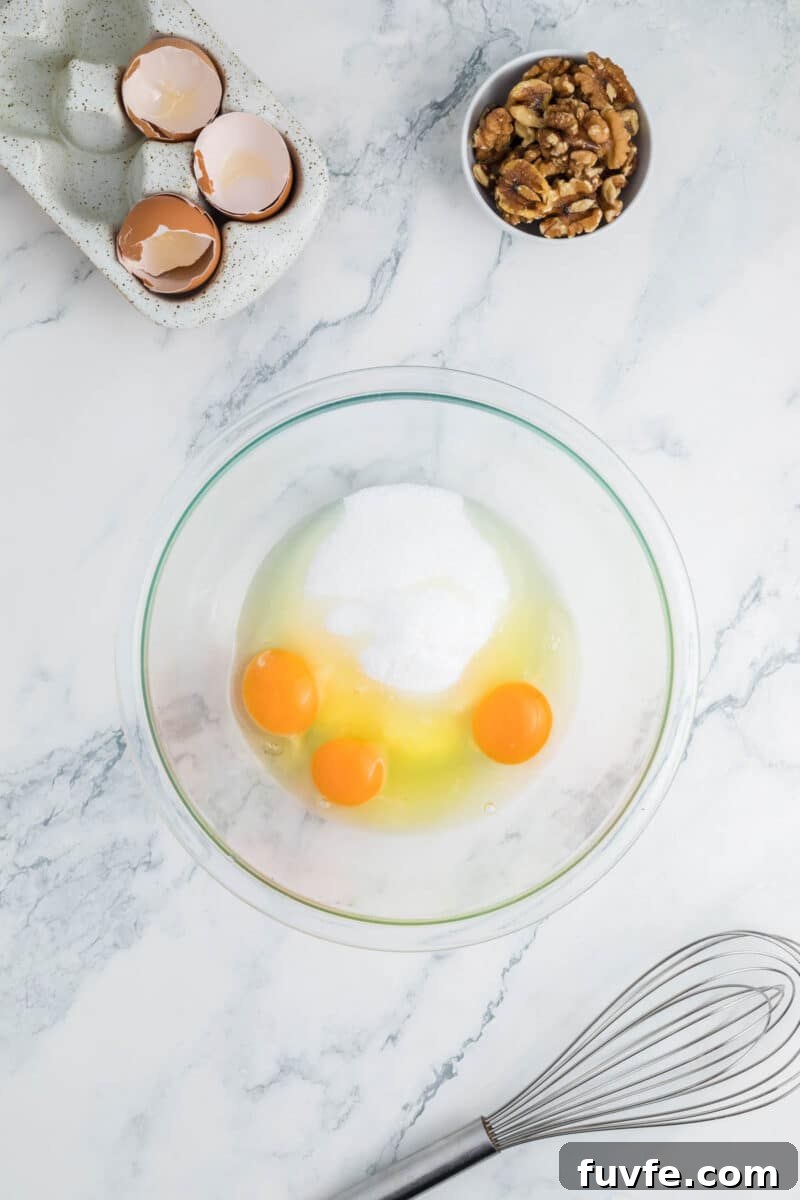 A glass bowl with three eggs and sugar, a whisk, a small bowl of walnuts, and cracked eggshells on a marble countertop.