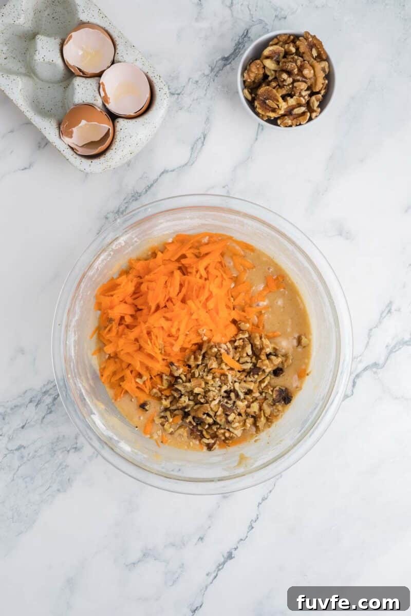 A glass bowl with cake batter, shredded carrots, and chopped walnuts sits on a marble countertop.