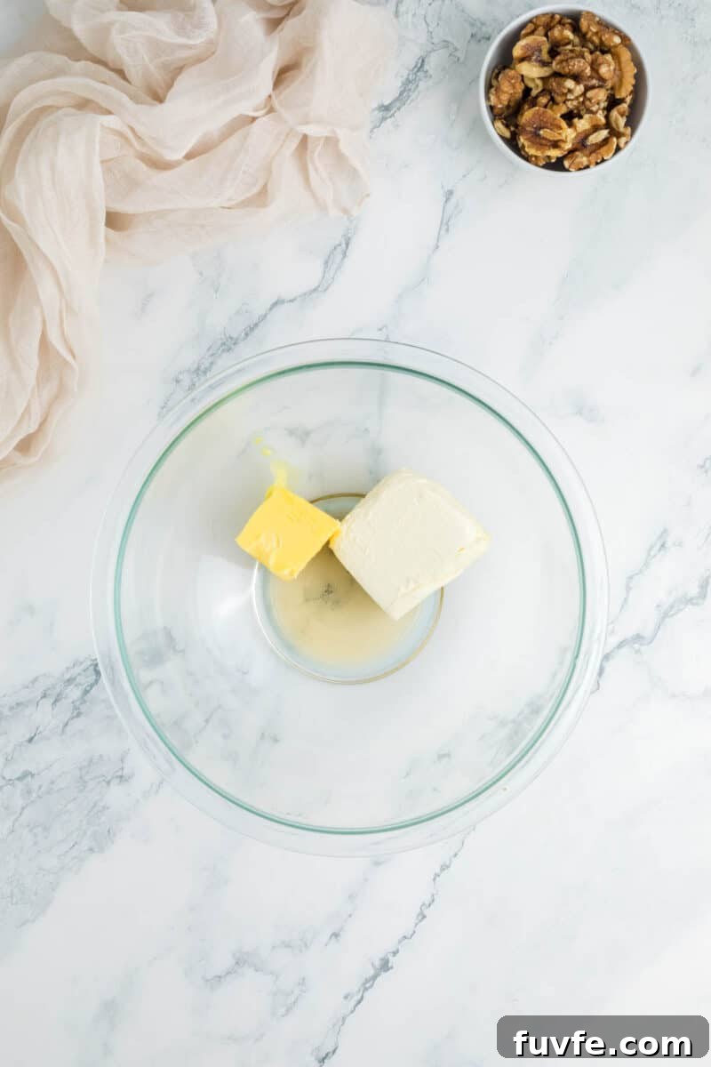 A glass bowl with a block of cream cheese and a chunk of butter on a marble surface.