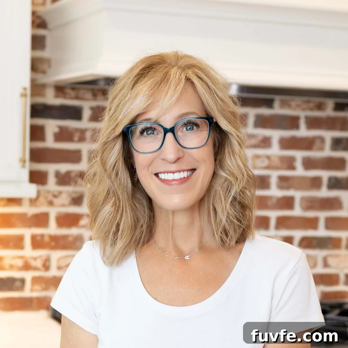 Person with shoulder-length blond hair and blue glasses smiling indoors in front of a red brick wall.