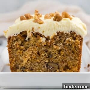 A sliced loaf of carrot cake with cream cheese frosting and chopped walnuts on top, displayed on a white dish.