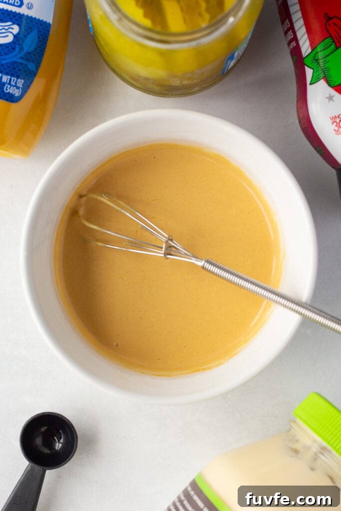 Quick 4-Ingredient Burger Sauce Recipe for Classic Flavor 6 Top down shot of a golden burger sauce in a small white dish with a small whisk in it, surrounded by jars and bottles of mayo, ketchup, yellow mustard, and dill pickles.