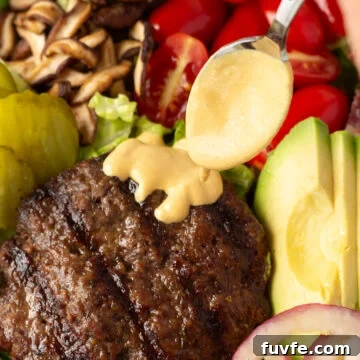 Quick 4-Ingredient Burger Sauce Recipe for Classic Flavor 8 Close up of a golden burger sauce being spooned on top of a burger patty on a salad with mushrooms, tomatoes, avocado, onions, and pickles on it.