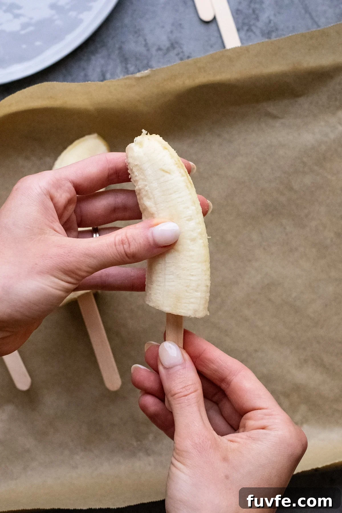 hand placing half of banana onto popsicle stick with parchment paper in the background.