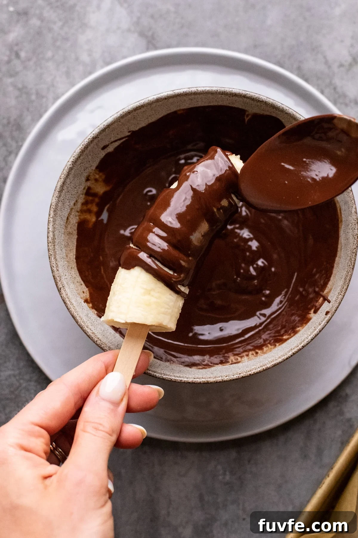 hand holding popsicle stick with banana and drizzling melted chocolate.