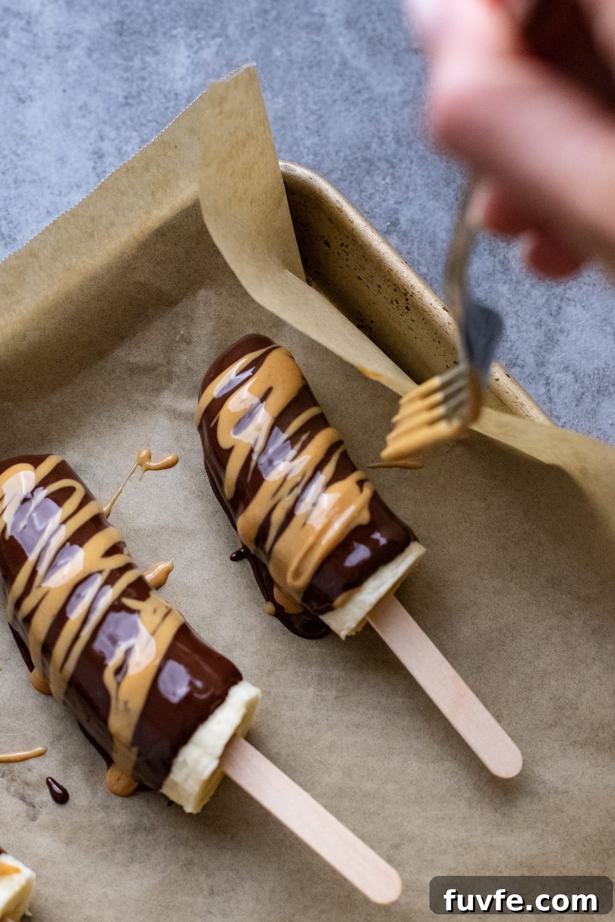 fork drizzling peanut butter over chocolate covered bananas on pan lined with parchment paper.