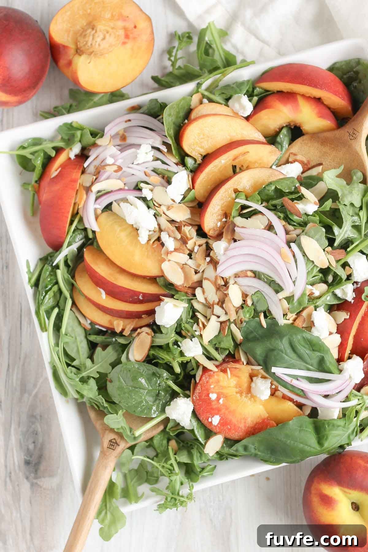 Wooden salad tongs mixing together a peach and goat cheese salad on a square plate with peaches on the sides.