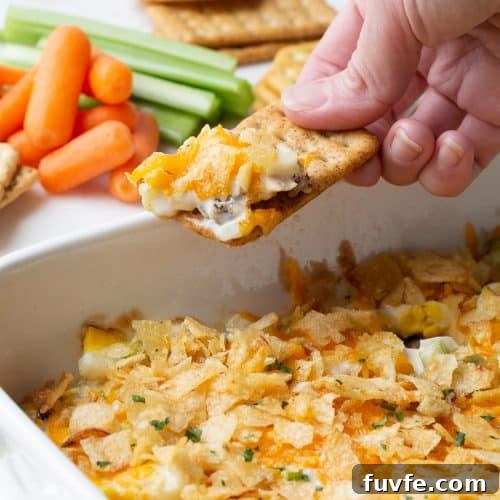 hand dipping a cracker into hot chicken salad topped with potato chips.