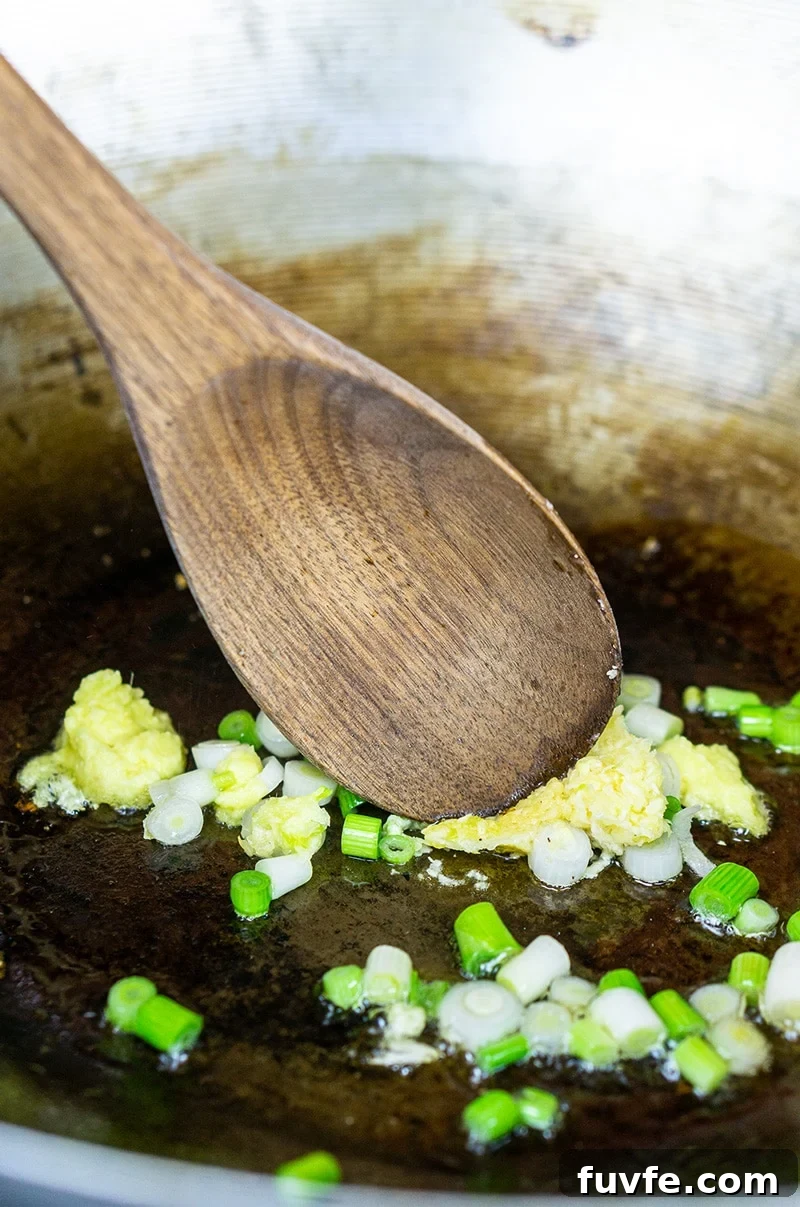 Garlic, ginger, oil, and green onions being sauteed in a skillet with a wooden spoon.