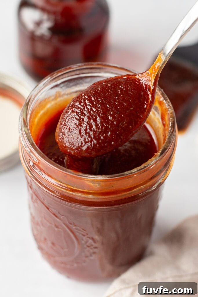 a small mason jar filled with maple bbq sauce that has a spoon bringing sauce out of it. The jar is on a white surface and in the background is a lid, a small jar of maple syrup, and a brush with bbq sauce on it.
