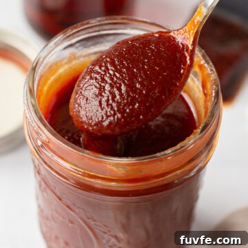 a small mason jar filled with maple bbq sauce that has a spoon bringing sauce out of it. The jar is on a white surface and in the background is a lid, a small jar of maple syrup, and a brush with bbq sauce on it.