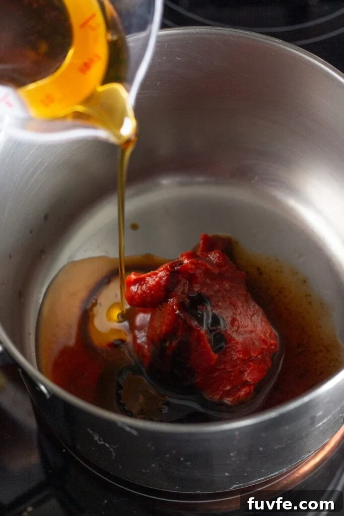 pouring maple syrup into a small saucepan on the stove that has tomato paste in it.