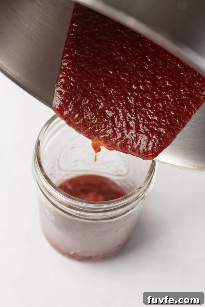 Close up of bbq sauce being poured out of a sauce pan and into a small mason jar on a white surface.