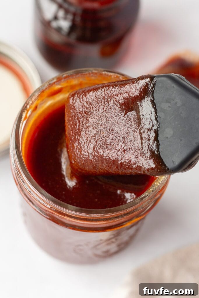 Close up of a basting brush being dipped into a small mason jar of bbq sauce that's resting on a white surface. In the background is a lid, a small container of maple syrup, a tan cloth napkin, and a spoon with bbq sauce on it.