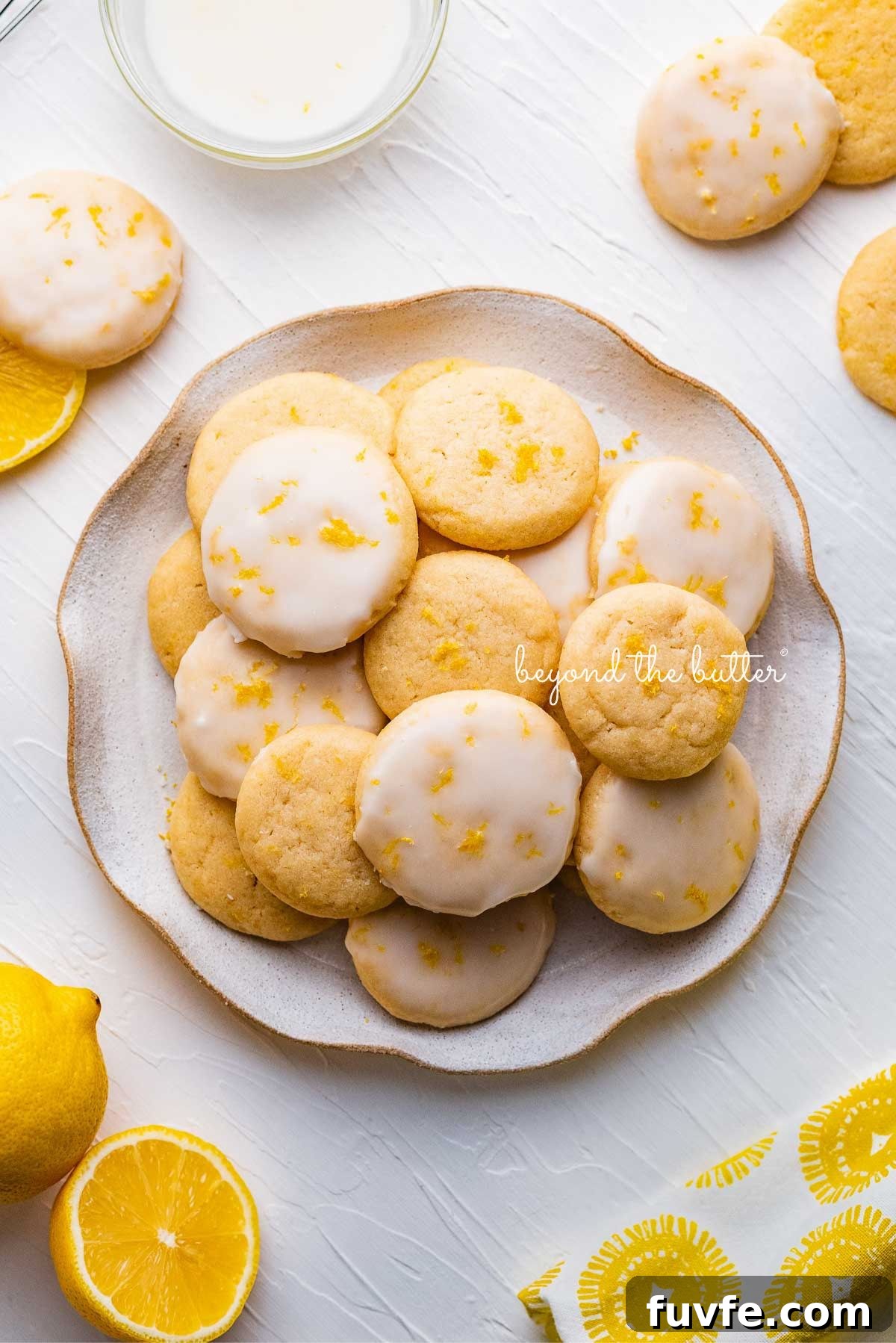 Plate full of lemon shortbread cookies surrounded by a lemon themed cloth napkin, lemons, and glass of milk on a white background.
