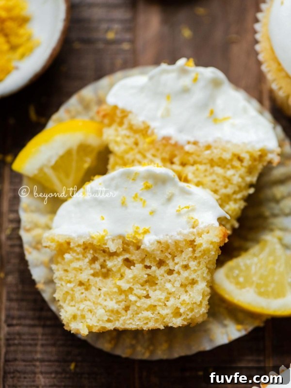 Sliced open lemon cupcake with whipped lemon cream cheese frosting on cupake liner and 2 wedges of lemons on a dark wood background.
