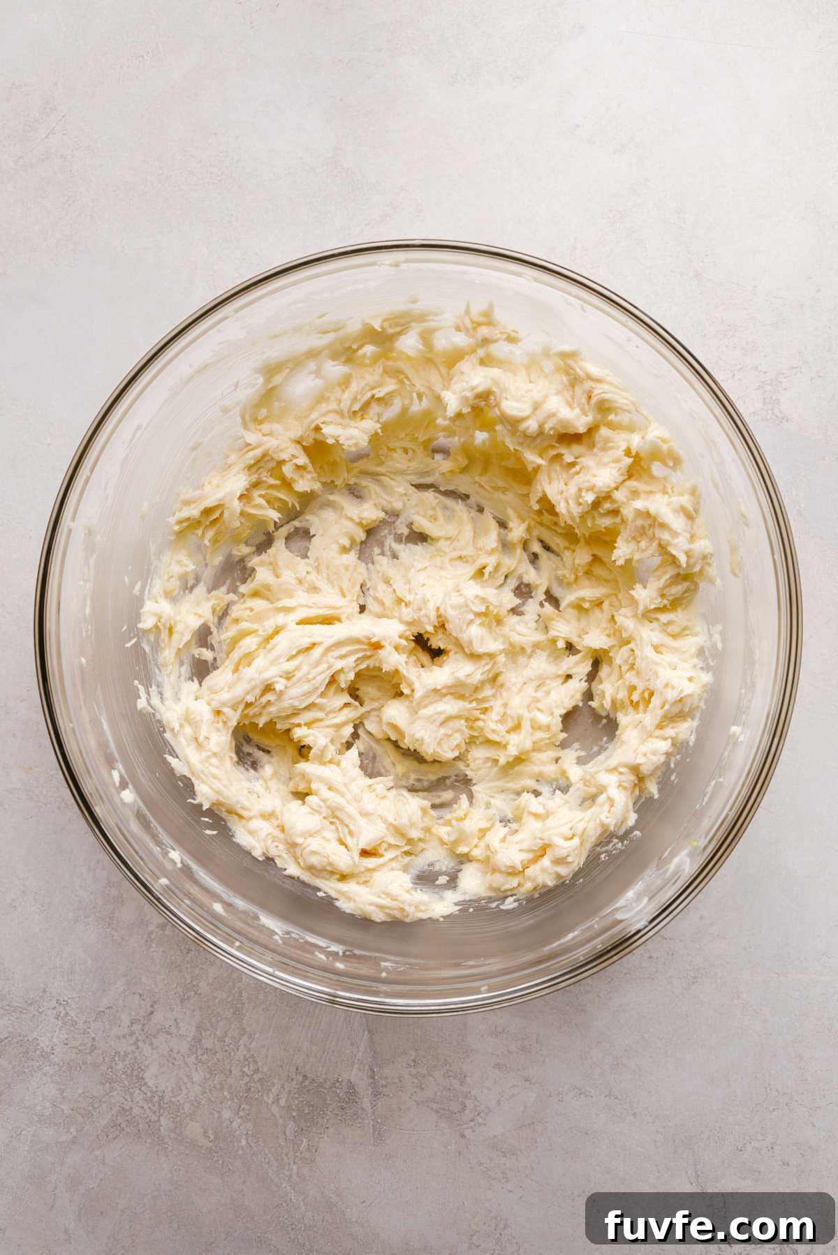 Whisked unsalted butter, granulated sugar, light brown sugar, lemon zest, lemon juice, and vanilla extract in a large glass mixing bowl on a grey marbled background.