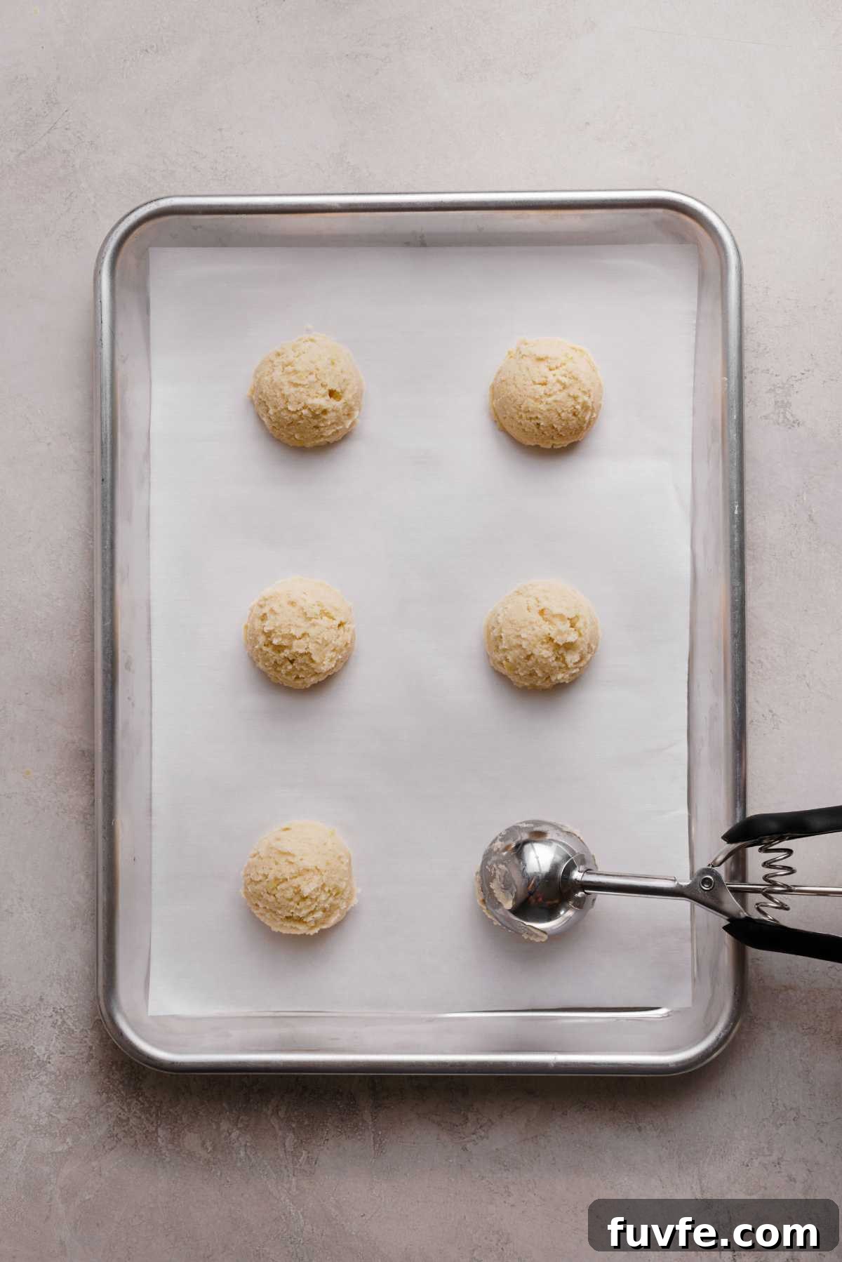 Scoops of lemon shortbread cookie dough on a white parchment paper lined baking sheet using a cookie scoop.