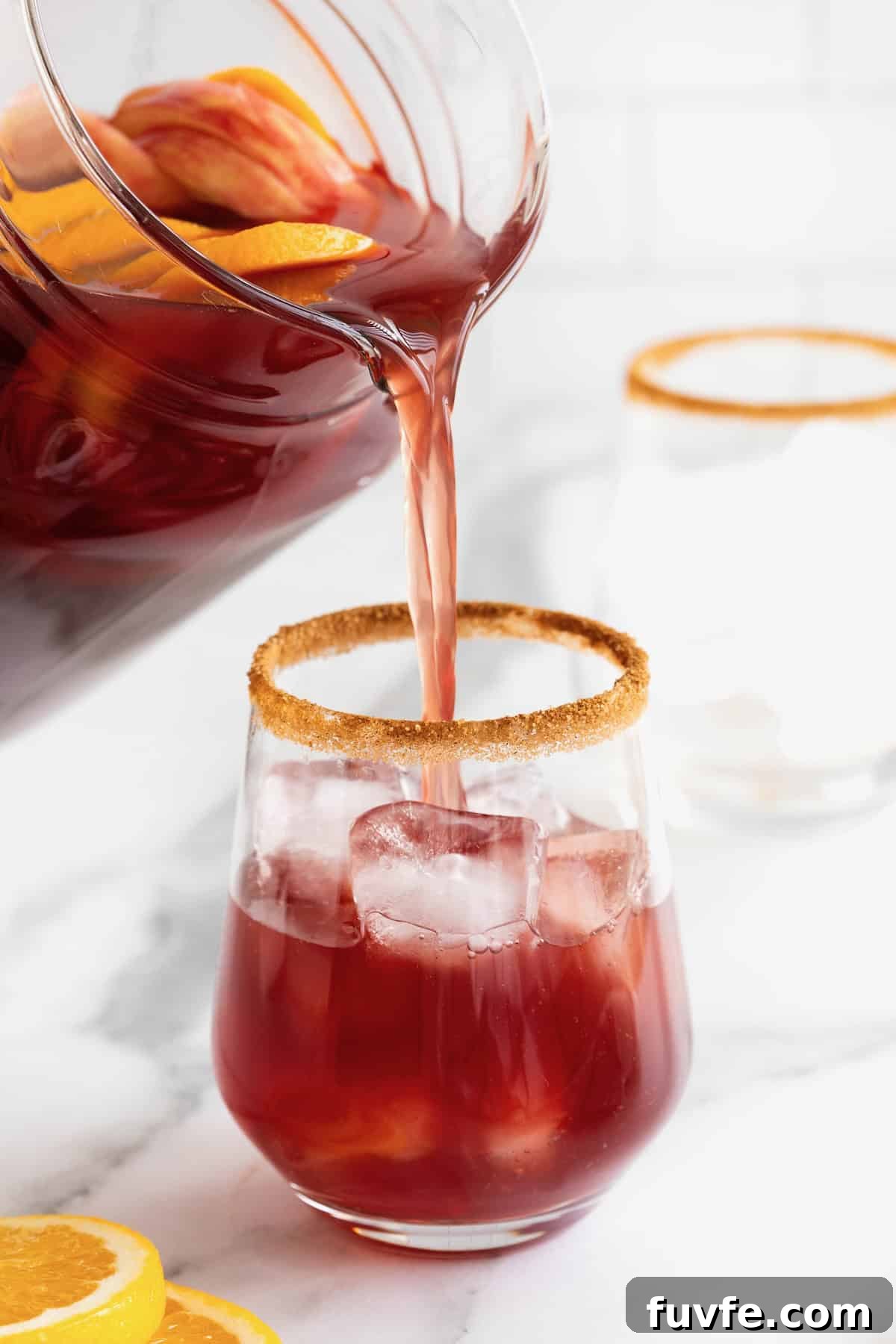 Apple cider sangria being poured from a pitcher into a cinnamon sugar rimmed glass with ice.