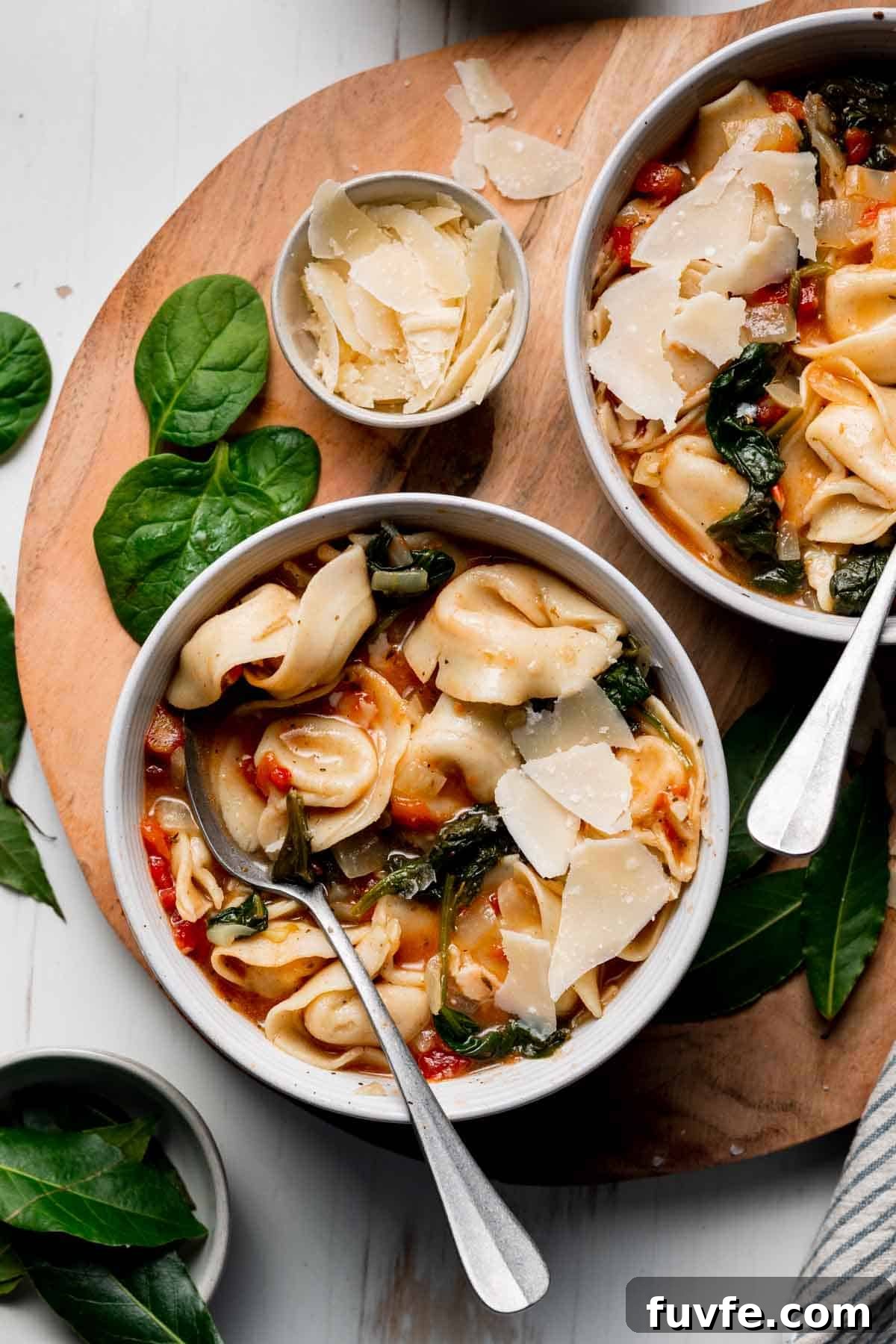 Two bowls of gluten free tortellini soup next to shaved parmesan and baby spinach on a wooden surface.