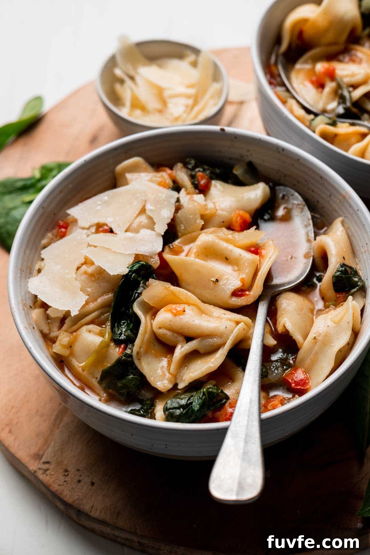 Tortellini soup in bowl with spoon, spinach and parmesan cheese on top.