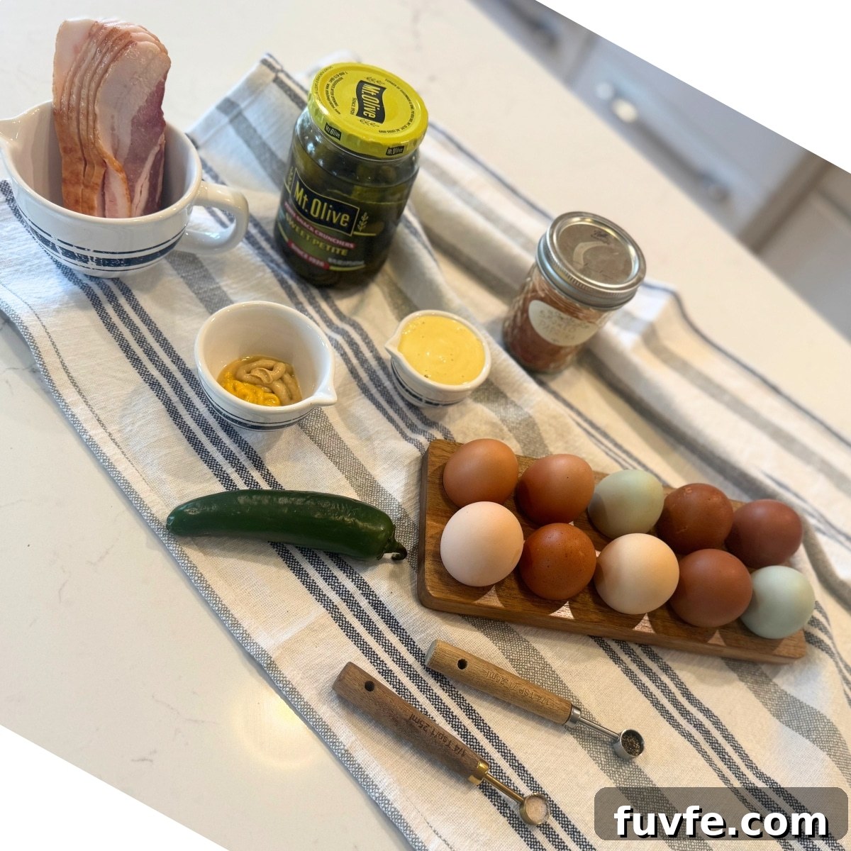 bacon jalapeno deviled eggs on counter: raw bacon, sweet pickled in a jar, smoked paprika in a jar, homemade mayo, yellow and dijon mustard in a dish, a jalapeno, salt, pepper and a dozen fresh eggs