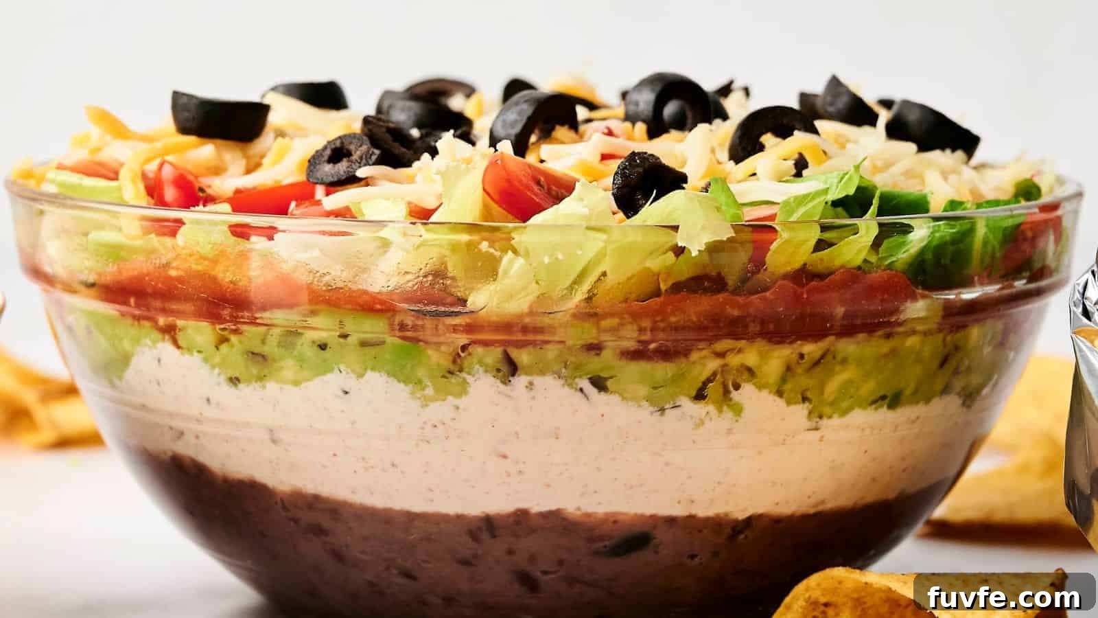 A clear glass bowl with 7 Layer Dip featuring beans, white cream, guacamole, diced tomatoes, shredded cheese, lettuce, and black olives.