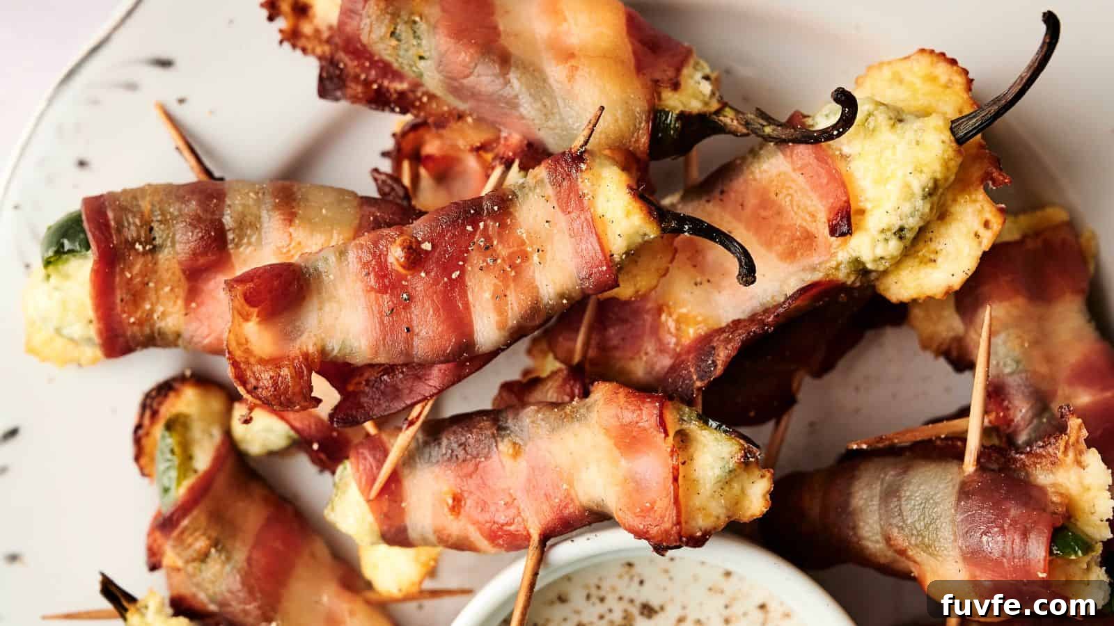 A plate of bacon-wrapped jalapeño poppers filled with cheese, secured with toothpicks, next to a small dish of dipping sauce.