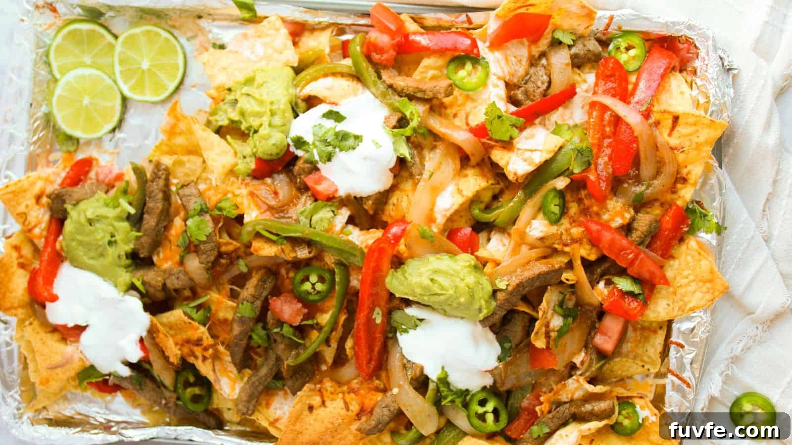 A tray of loaded nachos topped with sliced beef, red and green peppers, jalapeños, guacamole, sour cream, and lime wedges on the side.