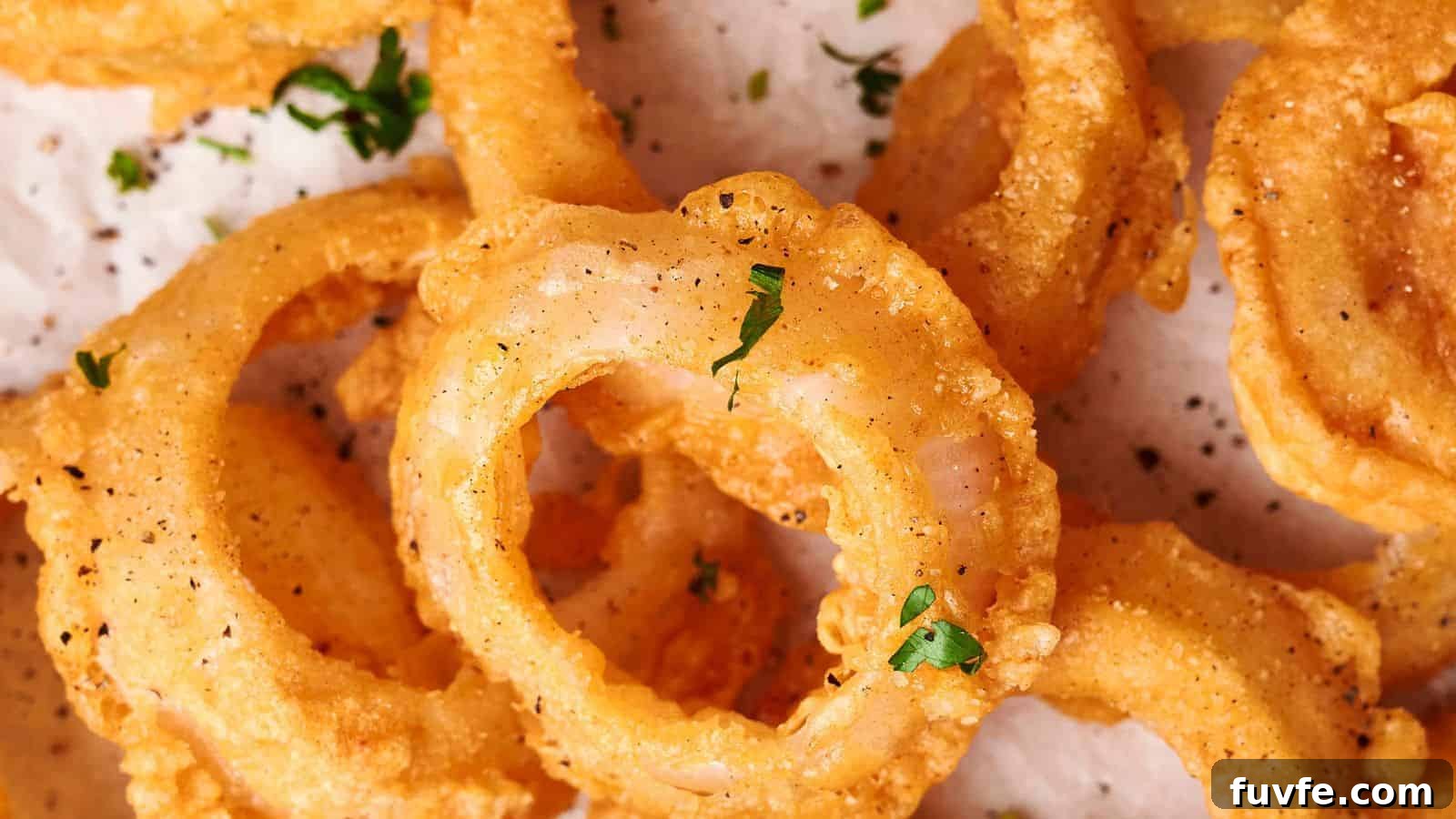 A close-up of crispy, golden-brown onion rings sprinkled with chopped parsley and black pepper on a white parchment-lined surface captures perfection in every bite.