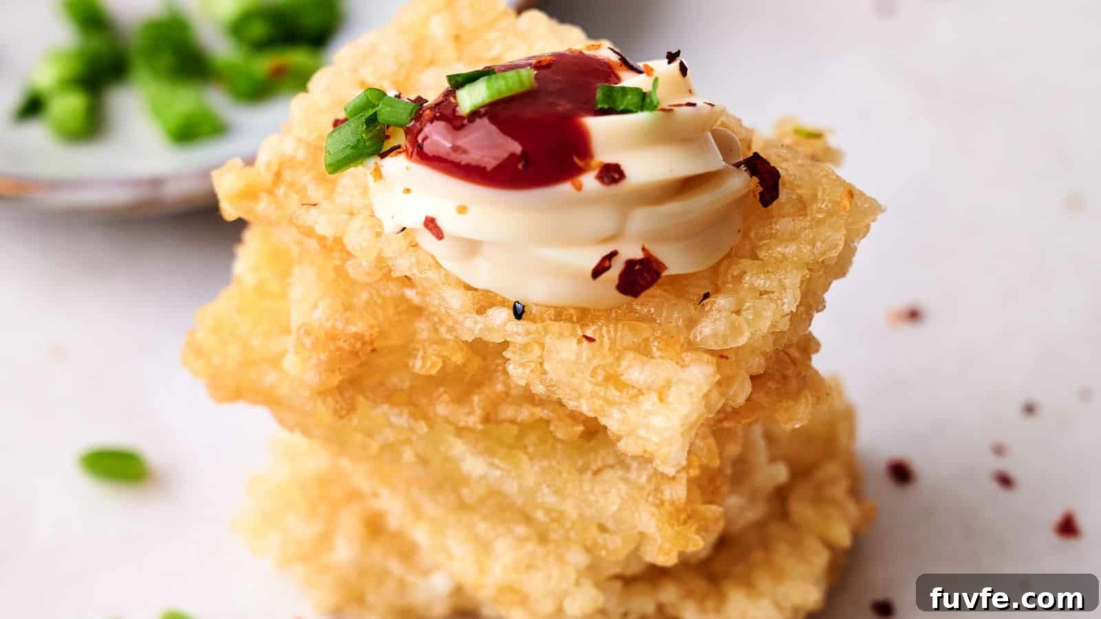 A stack of crispy rice topped with a dollop of white sauce, red sauce, and sliced green onions, with a blurred dish of green garnishes in the background.