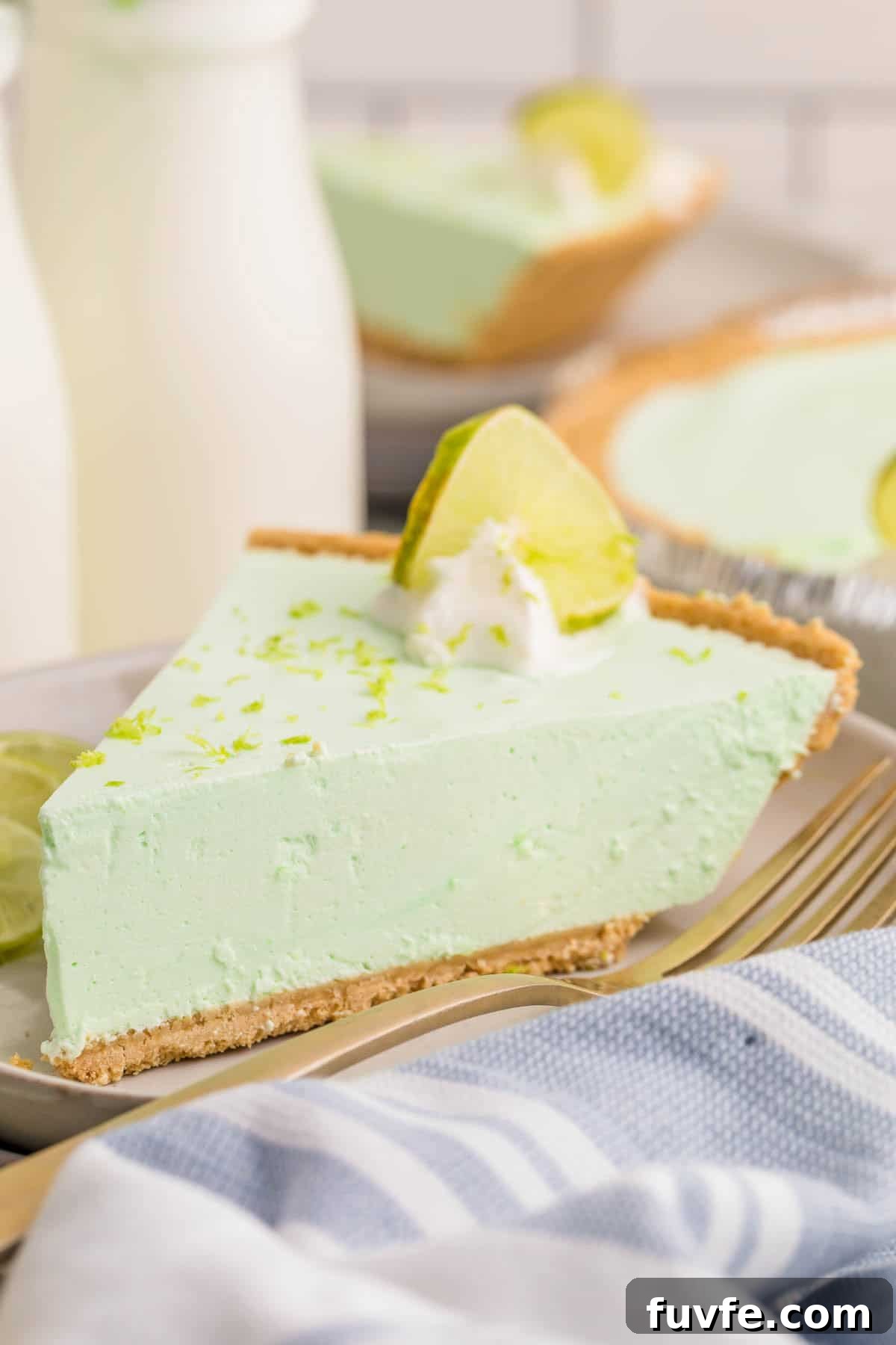 No-Bake Key Lime Jello Pie with Cool Whip Topping 2 Slice of key lime Jello pie with a graham cracker crust on a dessert plate. The pie is topped with whipped cream, a lime slice, and lime zest, with the remaining pie blurred in the background.