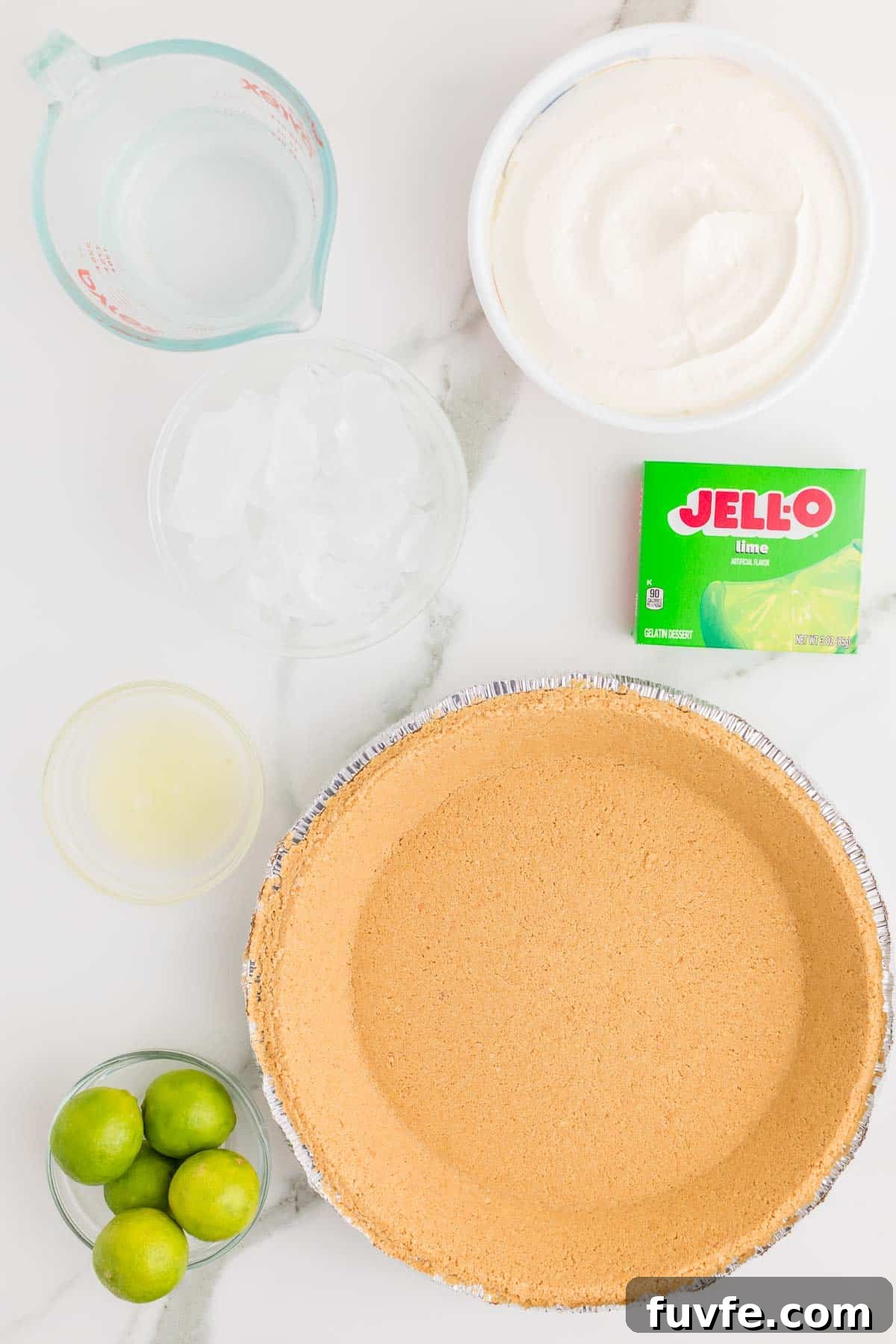 No-Bake Key Lime Jello Pie with Cool Whip Topping 3 Ingredients for key lime Jello pie arranged on a countertop. A prepared graham cracker crust sits alongside whipped topping, water, lime gelatin, lime juice, and whole limes. The gelatin package reads "JELL O Lime Artificial Flavor Gelatin Dessert."