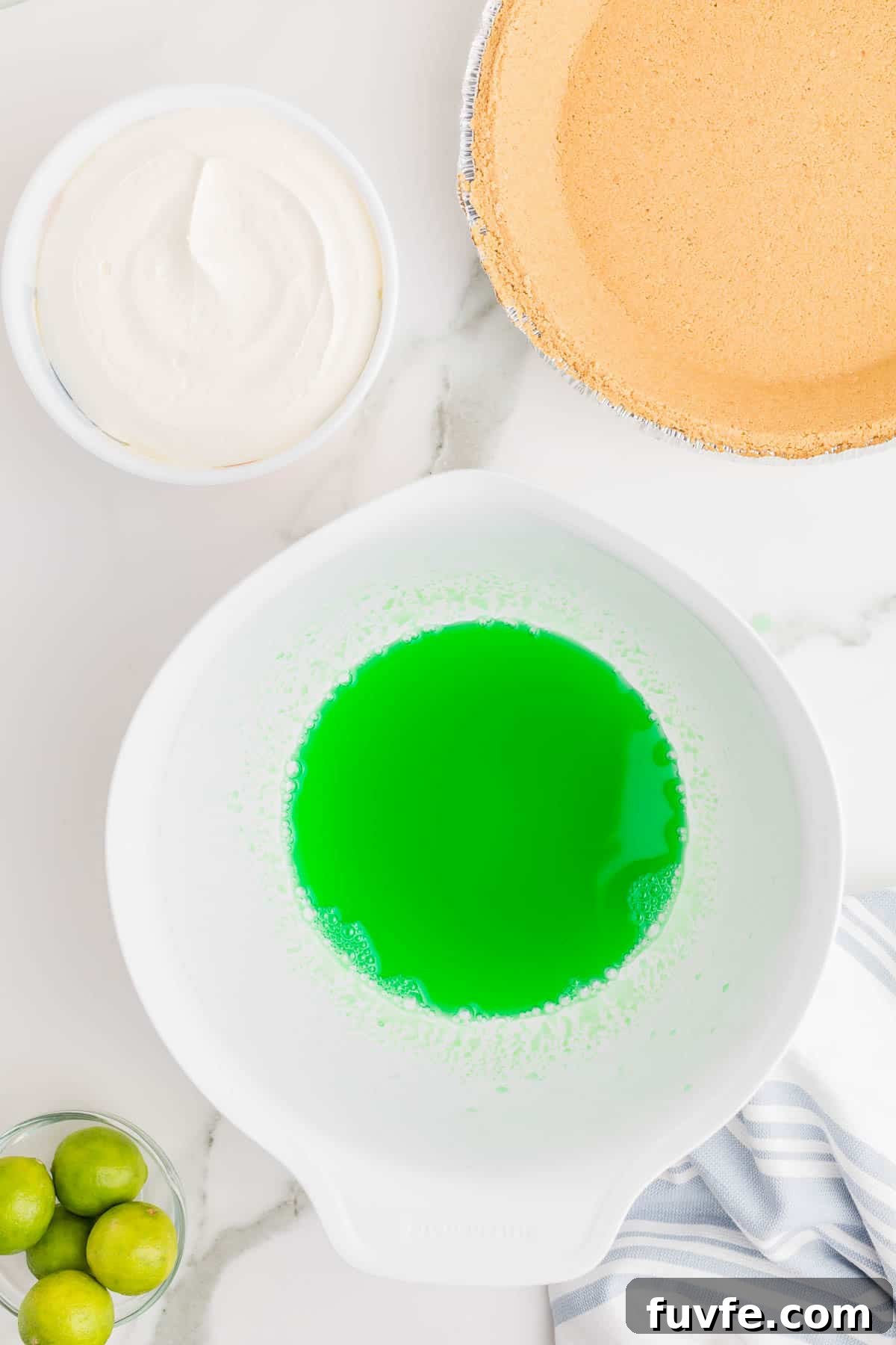 No-Bake Key Lime Jello Pie with Cool Whip Topping 4 Dissolved lime gelatin in a mixing bowl beside a graham cracker crust, whipped topping, and fresh limes. The mixture is prepared as the base for key lime Jello pie filling.