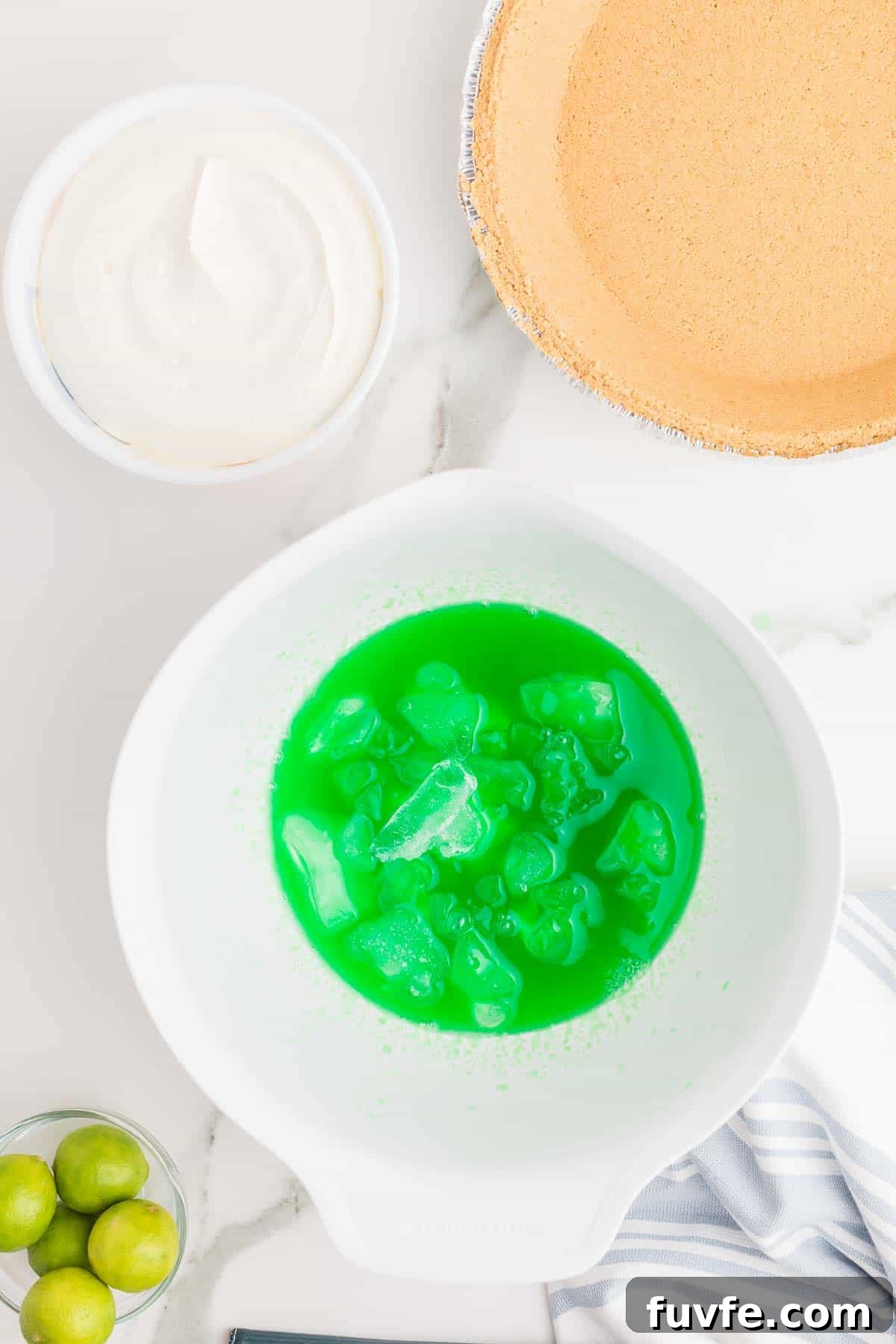 No-Bake Key Lime Jello Pie with Cool Whip Topping 5 Lime gelatin mixture in a bowl with ice cubes added to cool it before mixing. A prepared graham cracker crust and fresh limes are arranged nearby.